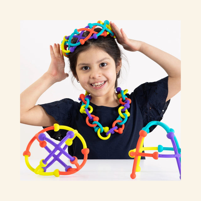 Child smiling while wearing colorful Fat Brain Toys Plip Kit pieces as a necklace and headband, with dome-shaped and arched Plip constructions displayed on the table.