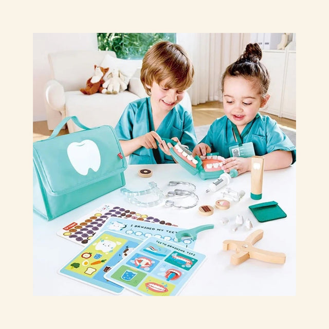 Two children playing with the Hape Super Smile Dental Clinic Set at a table, using the pretend dental tools, mouth model, activity cards, and turquoise carrying case.