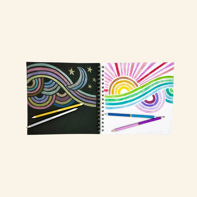 Artwork made with Snifty metallic colored pencils showing a rainbow design on black paper and a bright rainbow drawing on white paper with metallic pencils beside the sketchbooks.