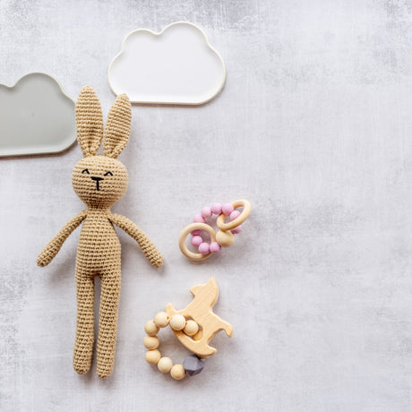 Flat lay of baby items on a soft gray textured background: a tan crocheted bunny with long ears, a pink and wood beaded teether ring, a wooden bead teether with a fox-shaped charm, and two cloud-shaped silicone dishes in white and sage green.