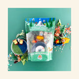 Camping KidDough Play Kit in green resealable pouch packaging on a dark green background, surrounded by kit contents: a jar of green and blue forest fir-scented play dough topped with log stampers, a teddy bear figurine in a yellow tent, a campfire, a forest park trail sign with a squirrel, miniature pine trees, and scattered pebble and campfire sprinkles.