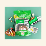 Earth Grown KidDoughs Garbage KidDough Play Kit shown with green retail bag packaging, brown root beer-scented dough, green garbage truck, raccoon figure, shovel, pretend trash pieces, banana peel, bottle, apple cores, and activity sheet arranged on a teal background.