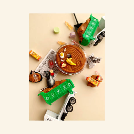 Earth Grown KidDoughs Garbage KidDough Play Kit shown from above with brown root beer-scented dough in a round container, green garbage truck, raccoon figure, shovel, pretend food scraps, bottles, bugs, and trash-themed accessories.
