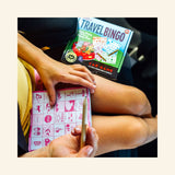 Child playing eeBoo Travel Bingo in the car, using a pencil to mark a pink bingo sheet while the Travel Bingo box sits on the seat nearby.