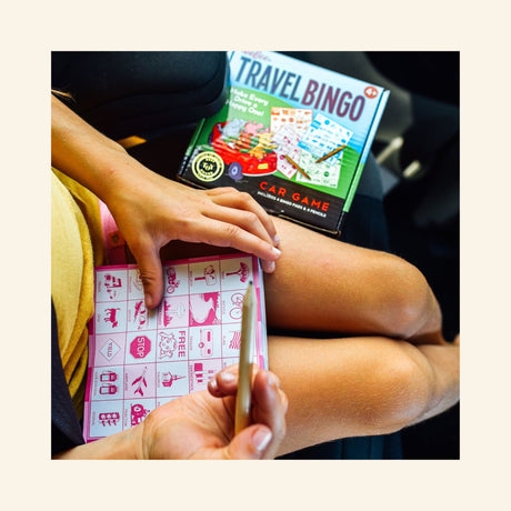 Child playing eeBoo Travel Bingo in the car, using a pencil to mark a pink bingo sheet while the Travel Bingo box sits on the seat nearby.