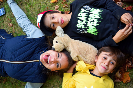 Two male children and one adult female lie together on the grass among fallen autumn leaves, smiling and making silly faces with a plush brown dog nestled between them.