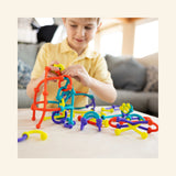 Child sitting at a table building with Fat Brain Toys Plip Kit pieces, with colorful connected silicone shapes and structures spread out in front of her.