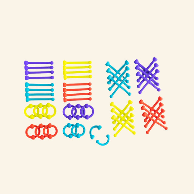 Fat Brain Toys Plip Kit pieces laid out by shape and color, showing silicone arches, sticks, ladder-like pieces, rings, and X-shaped connectors in red, yellow, blue, and purple.