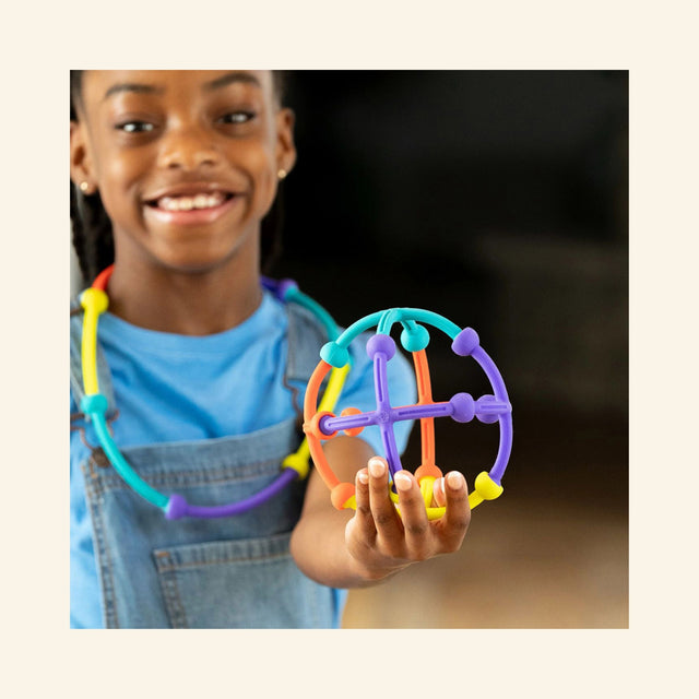 Child holding a round purple and orange Fat Brain Toys Plip Kit ball-like structure made from connected silicone arches and cross pieces.