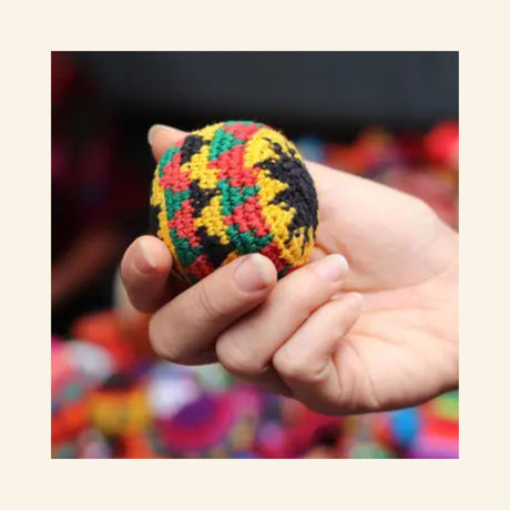 Hand holding a crocheted Guatemalan foot bag with a bright red, yellow, green, and black pattern against a colorful blurred background.