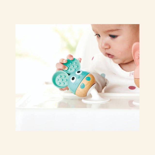 Baby playing with the teal mouse rattle from the Hape Cheery Critter Rattle Trio, holding the soft silicone ears while the rattle is attached to a smooth surface.