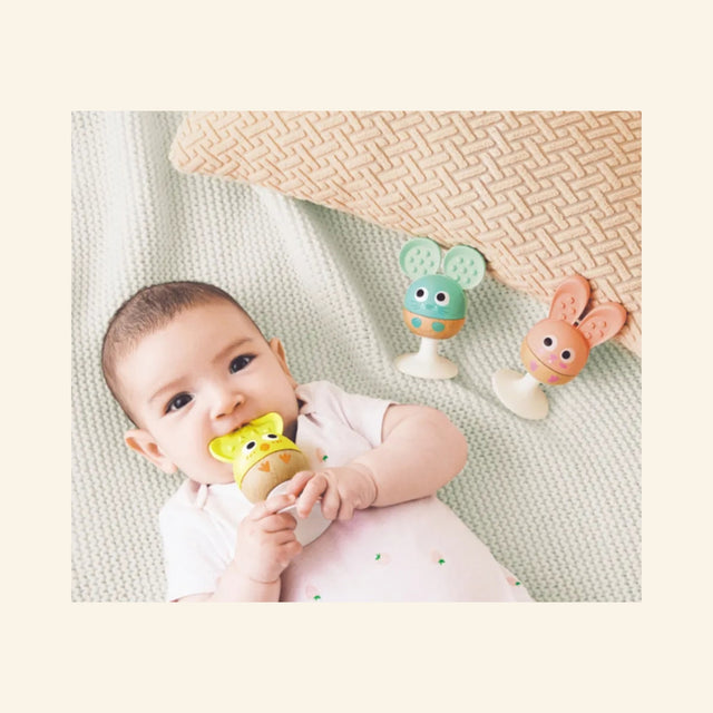Baby lying on a blanket with the Hape Cheery Critter Rattle Trio nearby, holding the yellow chick rattle while the mouse and rabbit rattles sit on their suction cup bases.