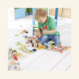 Child playing with the Hape Fix-It Tool Box on the floor, using a red hammer with wooden construction pieces, tools, gears, wheels, and bolts spread around for hands-on building play.