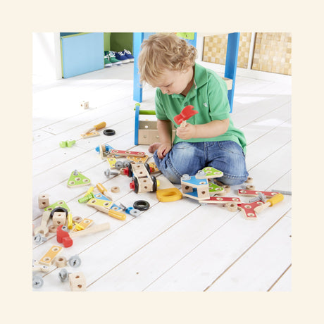 Child playing with the Hape Fix-It Tool Box on the floor, using a red hammer with wooden construction pieces, tools, gears, wheels, and bolts spread around for hands-on building play.