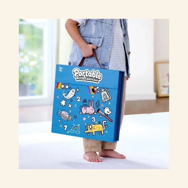 Child carrying the closed Hape Portable Art & Learn Set by the handle, showing the blue case decorated with rainbow, numbers, animals, art supplies, and playful learning graphics.