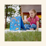 Child using the Hape Portable Art & Learn Set outdoors, drawing on the blue built-in easel with rainbow, number, animal, and space-themed artwork on the board.