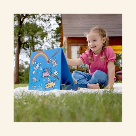 Child using the Hape Portable Art & Learn Set outdoors, drawing on the blue built-in easel with rainbow, number, animal, and space-themed artwork on the board.