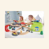 Two children playing with the Hape Railway Bucket Builder Set on a table, using the wooden train tracks, red bridge, green bucket hill, gray mountain lid, and crane accessories.