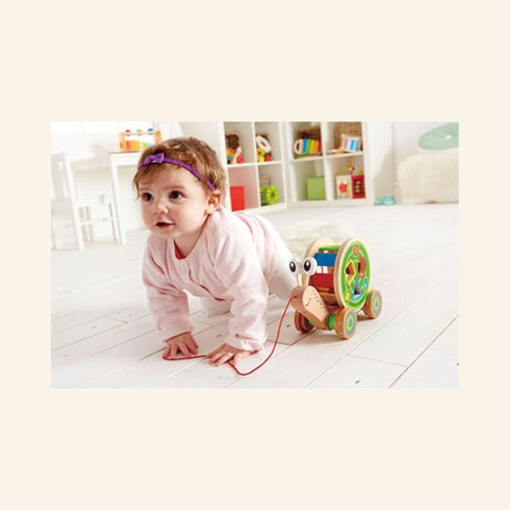 Baby crawling behind the Hape Walk-A-Long Snail on a white floor, using the red pull string while the wooden snail rolls ahead.