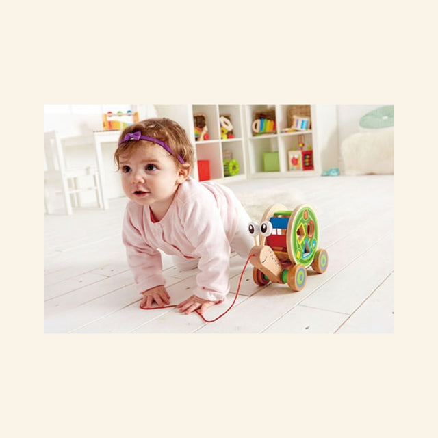 Baby crawling behind the Hape Walk-A-Long Snail on a white floor, using the red pull string while the wooden snail rolls ahead.