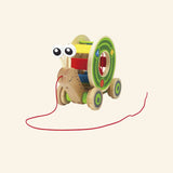 Hape Walk-A-Long Snail shown from the front with a wooden snail body, red pull string, colorful rolling shell, green wheels, and friendly eye stalks.