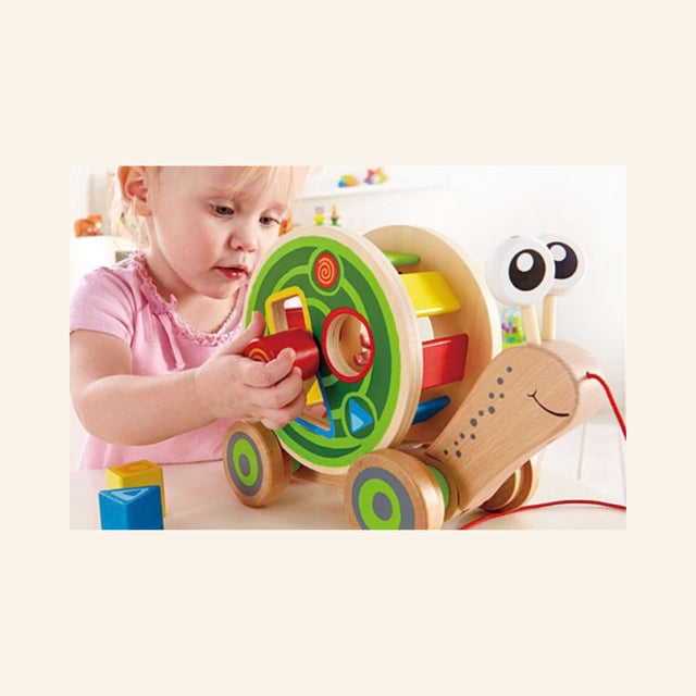 Toddler playing with the Hape Walk-A-Long Snail shape sorter, placing a red block into the colorful wooden snail shell with additional blocks nearby.