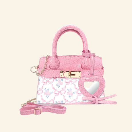 Hot Focus Charmie Luxe Crossbody – Bowtiful mini handbag shown in pink textured material with a bow-and-rose print front panel, gold-tone clasp and chain details, top handles, crossbody strap, and heart-shaped mirror charm.