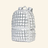 iScream Icy Silver Puffer Backpack shown from an angled side view, highlighting the metallic quilted puffer texture, side pocket, front zipper compartment, top handle, and padded shoulder straps.