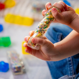 Child's hand holding a clear block that twists with colorful beads inside against a blurred background of toys.