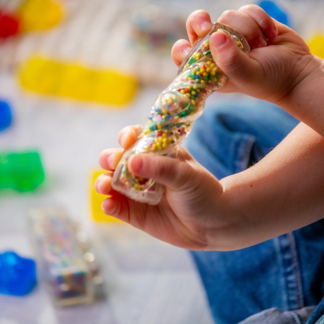 Child's hand holding a clear block that twists with colorful beads inside against a blurred background of toys.