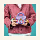 Child holding the LEGO® Creator Create Art Fashion Bag with Storage, showing the completed lavender mini handbag with colorful front tiles, handle, charms, and bow details.