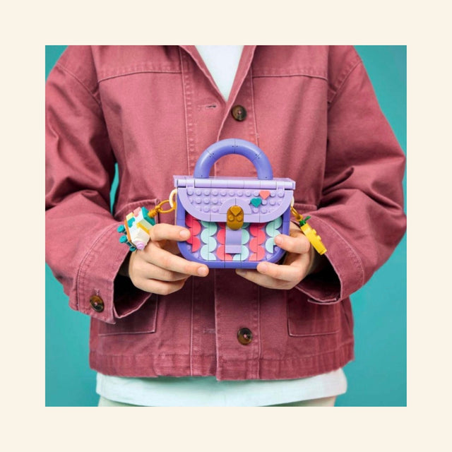 Child holding the LEGO® Creator Create Art Fashion Bag with Storage, showing the completed lavender mini handbag with colorful front tiles, handle, charms, and bow details.