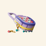 LEGO® Creator Create Art Fashion Bag shown at an angled top view with colorful pastel tile decorations, lavender handle, gold-style bow, and keychain charm pieces beside it.