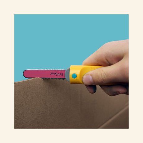 Hand using the yellow Makedo Safe-Saw to cut along a dotted line on cardboard, demonstrating the tool’s cardboard cutting function.