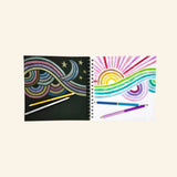 Artwork made with Snifty metallic colored pencils showing a rainbow design on black paper and a bright rainbow drawing on white paper with metallic pencils beside the sketchbooks.