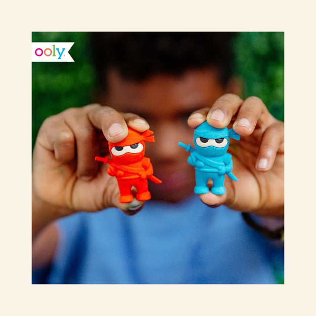 Child holding the red and blue OOLY Ninja Erasers, showing the colorful ninja-shaped erasers in use as playful school supplies.