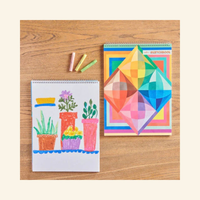 Rainbow Colors Sketchbook by eeBoo with colorful geometric cover beside an open sketchbook and oil pastels.