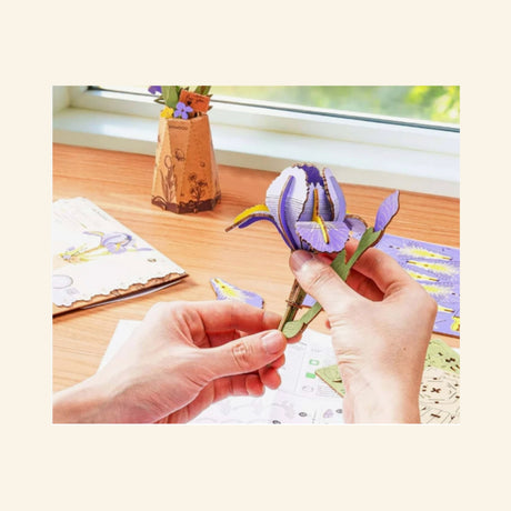 Hands assembling the Robotime Rowood Purple Iris wooden flower at a desk, with purple iris pieces, instruction sheets, and the finished vase displayed nearby.