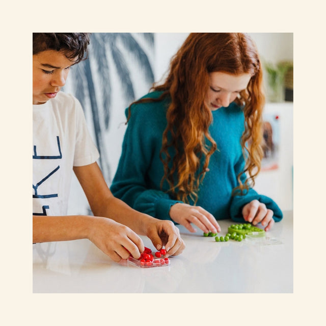 Two players using SmartGames IQ Mini puzzle games in red and green travel cases at a table, showing the compact game in use.