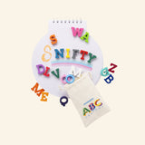 Snifty ABC Crayons spread around a small spiral notebook with colorful letter crayons spelling parts of words and a white printed storage pouch beside them.