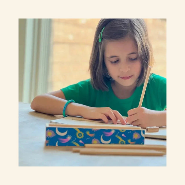Child sitting at a table drawing with a colored pencil, with the Snifty Cosmic Colored Pencil Box open nearby and natural wood colored pencils spread across the tabletop.