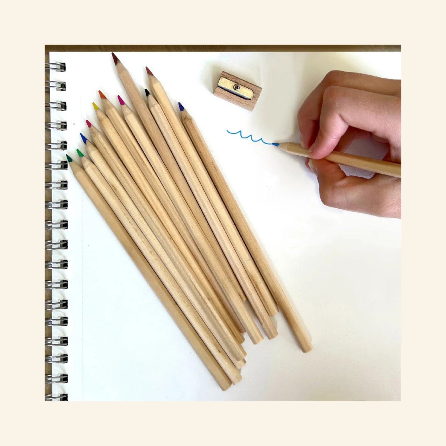 Snifty Cosmic Colored Pencil Box opened beside a spiral sketchbook, showing 12 natural wood colored pencils, a small wooden sharpener, and a hand drawing with a blue pencil.