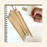 Snifty Cosmic Colored Pencil Box opened beside a spiral sketchbook, showing 12 natural wood colored pencils, a small wooden sharpener, and a hand drawing with a blue pencil.