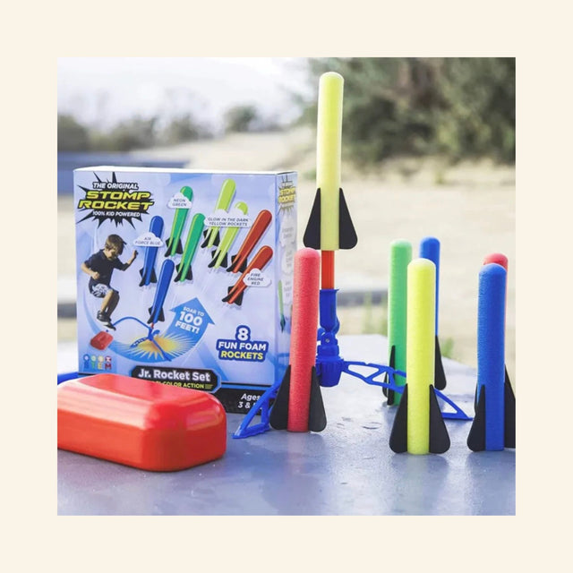 Stomp Rocket Jr. Rainbow Rocket Kit set up outdoors with colorful foam rockets standing beside the stomp launch pad, adjustable launcher, and product box.
