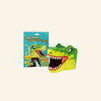 T-Rex Mask craft kit shown with teal packaging and a completed 3D paper T-Rex mask featuring green dinosaur details, big white teeth, expressive eyes, and open-mouth design.