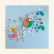 Flat lay of Unicorn KidDough Play Kit contents on a light blue background: an open jar of rainbow sherbet play dough topped with a pink unicorn figurine, a blue unicorn figurine, two silver star wands, rainbow and heart dough stampers, pastel star and heart molds, a clear rainbow cloud cutter, a mini corked jar of butterfly glitter, and a silver star garland with pastel confetti.