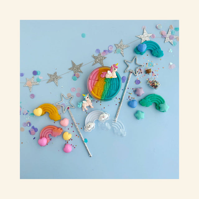 Flat lay of Unicorn KidDough Play Kit contents on a light blue background: an open jar of rainbow sherbet play dough topped with a pink unicorn figurine, a blue unicorn figurine, two silver star wands, rainbow and heart dough stampers, pastel star and heart molds, a clear rainbow cloud cutter, a mini corked jar of butterfly glitter, and a silver star garland with pastel confetti.
