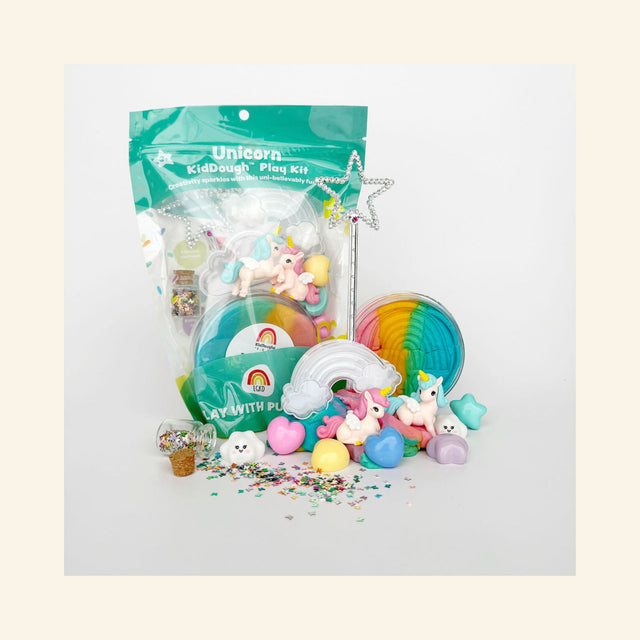 Unicorn KidDough Play Kit shown with its green resealable pouch packaging, surrounded by the kit's contents: a jar of rainbow sherbet play dough, two winged unicorn figurines, a silver star wand, a clear rainbow mold, pastel heart and star pieces, a smiling cloud, and a mini corked jar of glitter spilled in front.