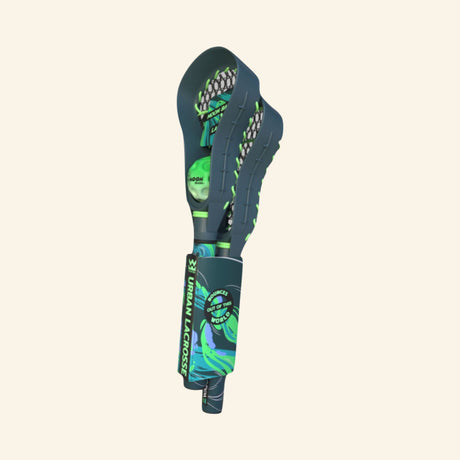 Side view of the Waboba Urban LAX mini lacrosse stick in retail packaging, showing the deep green head, netted pocket, blue-green handle graphics, and attached mini Moon Ball.