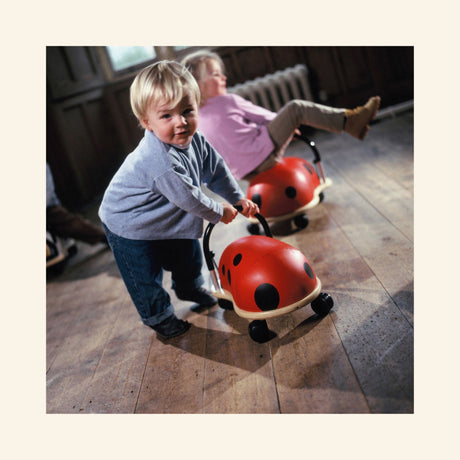 Toddler pushing and riding a red Wheely Bug Ladybug indoors while another child rides a second ladybug toy in the background on a hard floor.